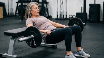 Una mujer de mediana edad con cabello grisáceo realiza un hip thrust con una barra cargada de pesas en un banco de gimnasio, vestida con ropa deportiva.