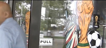 Primer plano de una puerta de cristal con un hombre de espaldas, calcomanías de la Estatua de la Libertad, la Copa Mundial de la FIFA y un balón de fútbol, y letreros 'PULL'