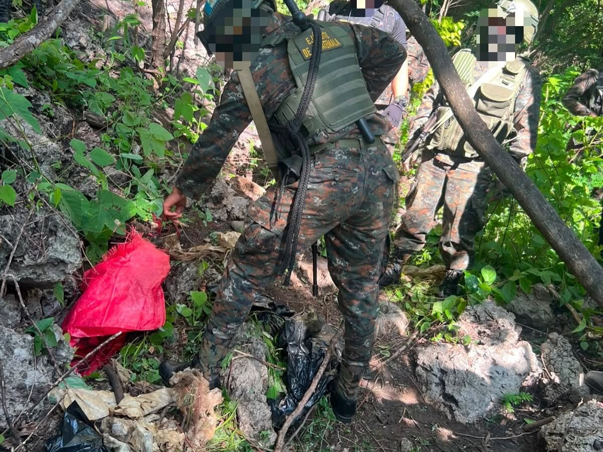 Un soldado del Ejército de Guatemala inspecciona el terreno cubierto de vegetación, junto a una bolsa plástica roja y otros elementos, durante una operación en una zona boscosa. (Ejército de Guatemala)
