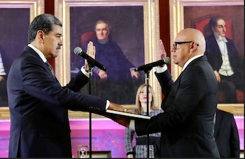 Dos hombres con traje, uno con bigote y otro calvo con gafas, se encuentran frente a micrófonos con las manos derechas levantadas. Uno sostiene un libro
