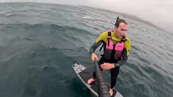 Primer plano de un hombre en traje de neopreno y chaleco rosado sobre una tabla de paddleboard en un mar agitado. Sostiene un palo. Otro navegante se ve a distancia