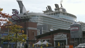 Un gran crucero blanco atracado junto a un paseo de madera donde caminan varias personas cerca de edificios y áreas verdes