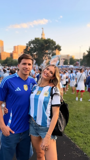 Eva Bargiela y Gianluca Simeone sonríen a cámara, él con camiseta azul de Argentina, ella con la albiceleste y maquillaje facial, en un parque lleno de fans