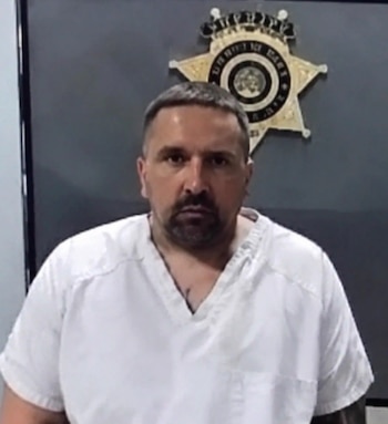 Retrato policial de un hombre con cabello corto, barba y camiseta blanca frente a una pared con una insignia dorada de la policía
