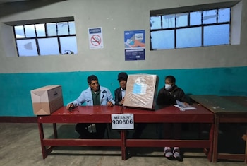 Três pessoas, uma delas usando máscara, sentam-se atrás de uma mesa de madeira vermelha em uma sala com paredes coloridas e verdes. Existem duas janelas e duas placas na parede. Sobre a mesa está a urna e a inscrição 'TABELA N° 900606'