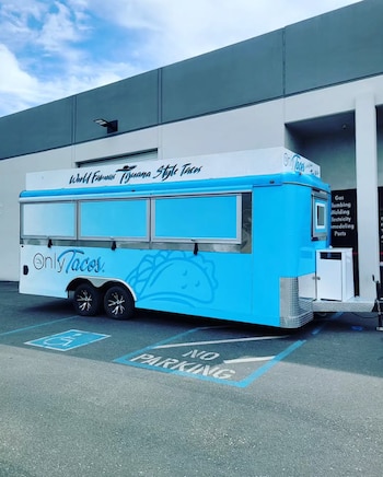 Remolque de comida de color azul y blanco con el texto Only Tacos estacionado frente a un edificio gris
