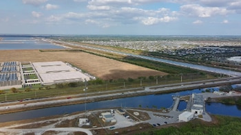 Vista aérea de grandes edificios industriales, paneles solares, una autopista con vehículos, un canal, una estación hídrica, campos y cuerpos de agua