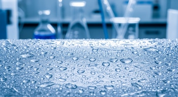 Primer plano de una superficie de material plástico o polímero translúcido cubierta con múltiples gotas de agua de distintos tamaños. Al fondo, equipo de laboratorio desenfocado.