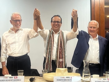 Roberto Sánchez Palomino, Ricardo Belmont Cassinelli y Alfonso López-Cha sonríen con las manos unidas. Mesa con placas, vasos y micrófonos