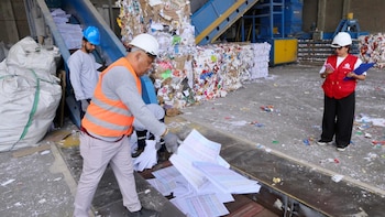 Un trabajador con chaleco de seguridad coloca hojas de papel en una cinta transportadora ante la mirada de una supervisora con chaleco rojo