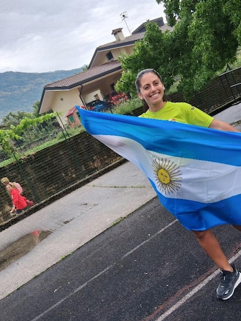 Una mujer con camiseta verde sostiene la bandera extendida de Argentina en una vía asfaltada junto a una cerca y una vivienda