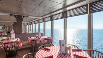Restaurante con mesas de cuadros rojos y blancos, sillas de madera y bancos. Grandes ventanas con persianas superiores revelan el vasto océano brillando bajo el sol