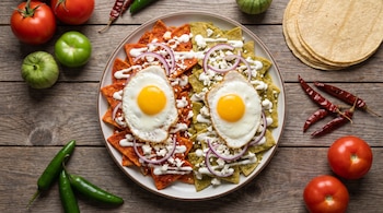 Vista cenital de un plato con chilaquiles rojos y verdes, huevos fritos, queso, crema y cebolla, rodeado de tomates, chiles y tortillas.