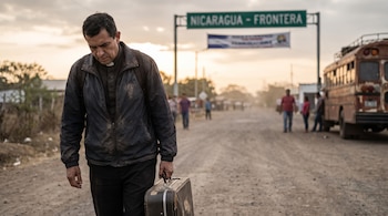 Un hombre con cuello clerical y chaqueta oscura camina en un camino de tierra con una maleta. Al fondo, un letrero de "Nicaragua - Frontera", personas y un autobús.