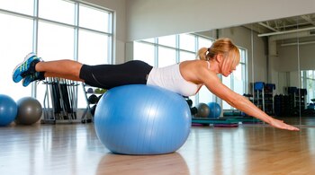 Mujer rubia en top blanco y shorts negros realiza un ejercicio de equilibrio sobre un balón de estabilidad azul en un gimnasio con ventanas y espejos.