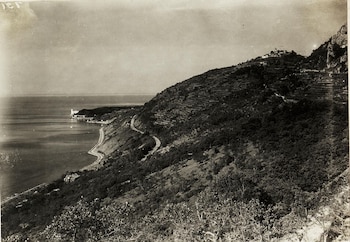 Foto monocromática de una costa con mar y colinas. Un camino serpentea la ladera boscosa, con terrazas y edificios. Un pueblo se asienta en la cima