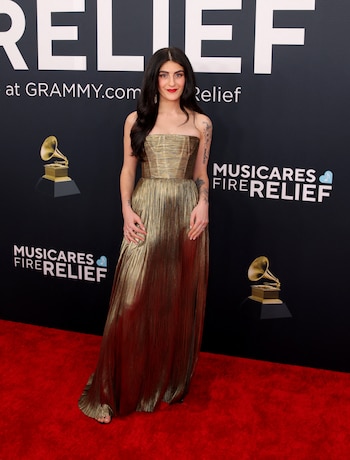 La compositora Kara Talve llegó con un vestido largo en tonos dorados con un acabado metálico y un diseño sin mangas. La prenda presenta un corte ajustado en la parte superior y una caída fluida hasta el suelo, con pliegues que crean un efecto visual brillante bajo la iluminación