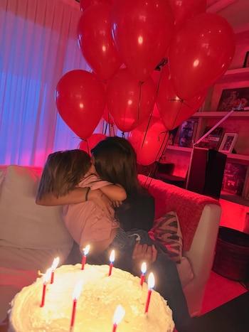 Calu Rivero abraza a Aito de la Rua en un sofá, con globos rojos al fondo y un pastel blanco con velas encendidas en primer plano