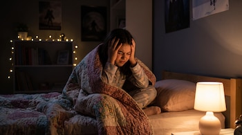 Una niña en pijama sentada en la cama en una habitación oscura con lámpara y luces decorativas. Se agarra la cabeza con una mueca de dolor.