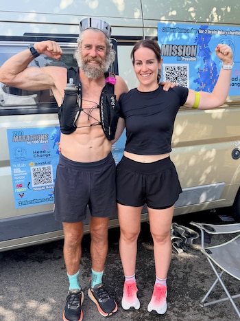 Un hombre y una mujer con ropa deportiva flexionan los brazos junto a un vehículo con carteles informativos sobre un reto maratónico