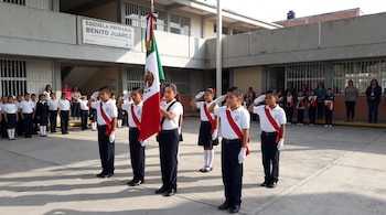 Niños de escolta con uniforme, banda roja y guantes blancos rinden honores a la bandera de México. Fondo con edificio escolar y más alumnos.
