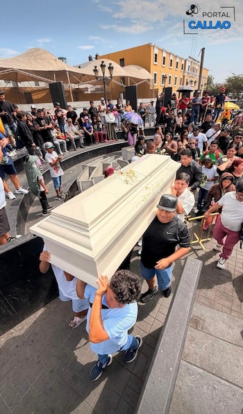 Un féretro blanco es llevado por varias personas, seguido por una gran multitud en un espacio público al aire libre con edificios amarillos de fondo