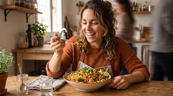 Una mujer sentada a una mesa de madera sostiene una cuchara con comida frente a un tazón de arroz, garbanzos y verduras.