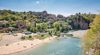 Vista de casas de piedra en una colina verde junto a un río, una orilla de arena con bañistas y un aparcamiento de vehículos.