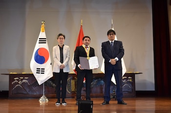 Tres personas posan en un escenario frente a las banderas de Corea del Sur y Perú mientras la del centro sostiene una medalla y un diploma