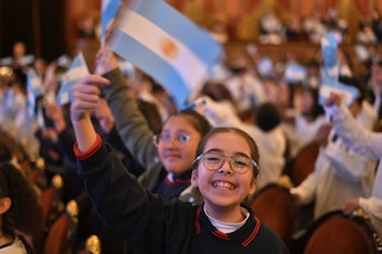 Una niña con gafas en primer plano levanta un brazo. En el fondo se observan otras personas, varias banderas argentinas y butacas de un auditorio