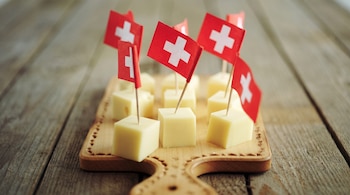 Una tabla de madera contiene cubos de queso pálido con palillos que tienen banderas de Suiza en la parte superior.