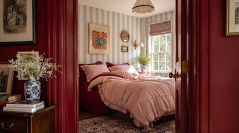 Un dormitorio con cama vestida, paredes a rayas azules y blancas, un póster de Matisse, una mesita de noche con un jarrón de flores y libros.