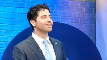 Plano medio de Manny Rutinel con traje azul, camisa blanca y corbata clara, sonriendo frente a un fondo azul con textura de ladrillo