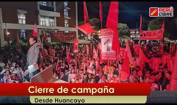 Hombre en un escenario hablando a una gran multitud de noche, rodeado de numerosas banderas rojas y pancartas con mensajes políticos