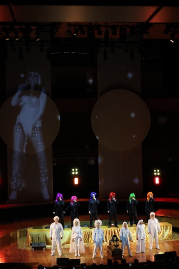 Escenario con artistas. Siete vestidos de blanco con pelucas blancas, seis con trajes oscuros y pelucas de colores. Proyecciones de siluetas y una estatua dorada