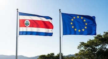 Dos astas de bandera con la bandera de Costa Rica a la izquierda y la bandera de la Unión Europea a la derecha, ondeando bajo un cielo azul claro.