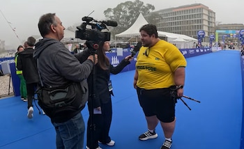 Camerógrafo filma a reportera entrevistando a hombre con camiseta amarilla de Brasil en pista azul. Toldo blanco y edificios se ven al fondo, día nublado