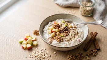 Tazón con yogur griego, trozos de manzana, nueces, avena y canela sobre una superficie de madera con ingredientes dispersos alrededor.