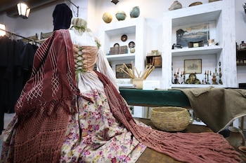 Un vestido de época sobre un maniquí con chal rojo y falda floral, junto a una cesta de mimbre y estantes con libros y globos terráqueos