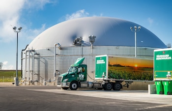 Gran domo gris de planta de biogás con tuberías, junto a un camión verde con remolque de Vanguard Renewables. Campo verde y cielo azul de fondo