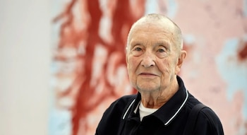 Primer plano de Georg Baselitz, un hombre mayor de piel clara y cabeza afeitada, vestido con polo negro, sobre un fondo difuso de una pintura abstracta