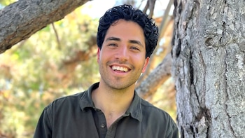 Un hombre con cabello oscuro rizado y camisa verde oscuro sonríe junto al tronco de un árbol
