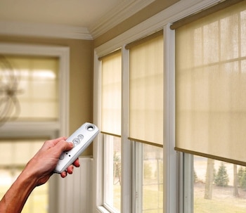 Una mano sostiene un control remoto blanco frente a tres ventanas con cortinas enrollables de color claro, parcialmente bajadas