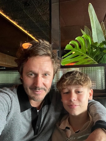 Un hombre adulto con barba y cabello castaño rizado posa junto a un adolescente de cabello rubio, ambos sonríen levemente a la cámara. De fondo, plantas