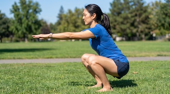 Mujer de perfil con cabello oscuro recogido en coleta, camiseta azul y shorts oscuros, descalza sobre césped verde, realizando una sentadilla con brazos extendidos.