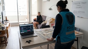 Una oficial de policía de espaldas, con chaleco azul, inspecciona una mesa con billetes de dólar, un portátil mostrando criptomonedas y llaves de auto.