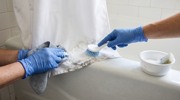 Manos con guantes azules frotan con cepillo y paño la parte inferior manchada de una cortina blanca junto a un recipiente con pasta.