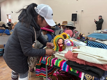 Mujer con gorra y chaqueta oscura organiza una mesa con textiles coloridos, muñecos de crochet (Virgen María y unicornio) y prendas de lana apiladas