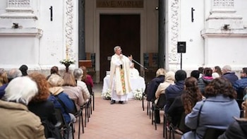 Jorge Garcia Cuerva, vestido de sacerdote, habla por micrófono a una congregación sentada al aire libre frente a la entrada blanca del Convento Santa Catalina de Siena