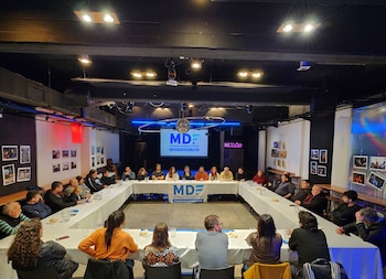 Grupo de personas sentadas alrededor de una mesa en forma de U; pantalla proyecta logo MDF y texto; banderas de MDF sobre la mesa; luces en el techo
